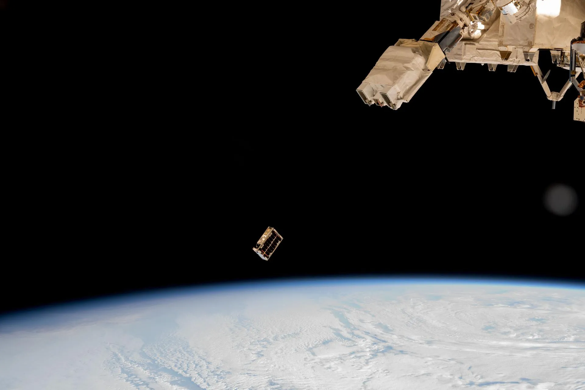 A small CubeSat drifts away from the space station's robotic arm, with the Earth's limb below